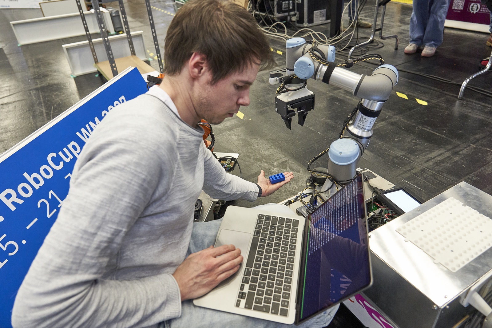 Ein Teammitglied mit Laptop und einem blauen Bauteil in der Hand programmiert den Roboterarm des mobilen Roboters