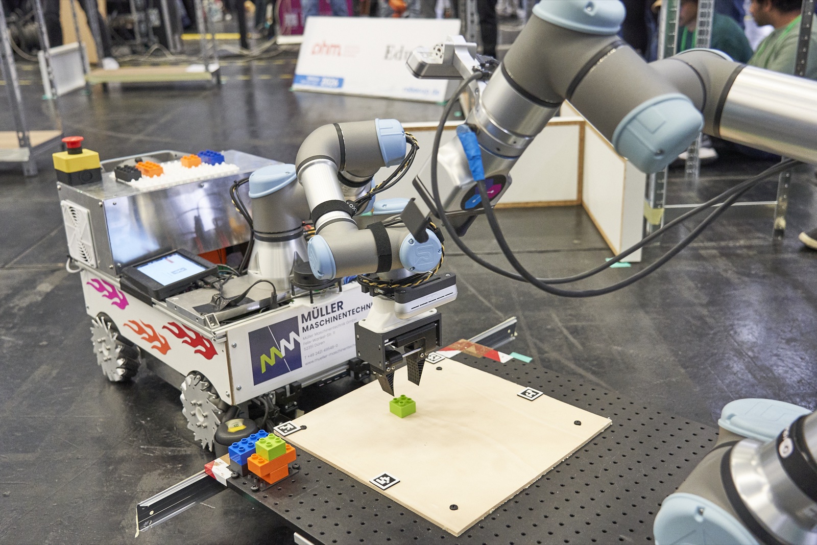 Der mobile Roboter von GM-Force Cologne mit Mecanum-Rädern und Roboterarm greift ein grünes Bauteil an einer Montagestation