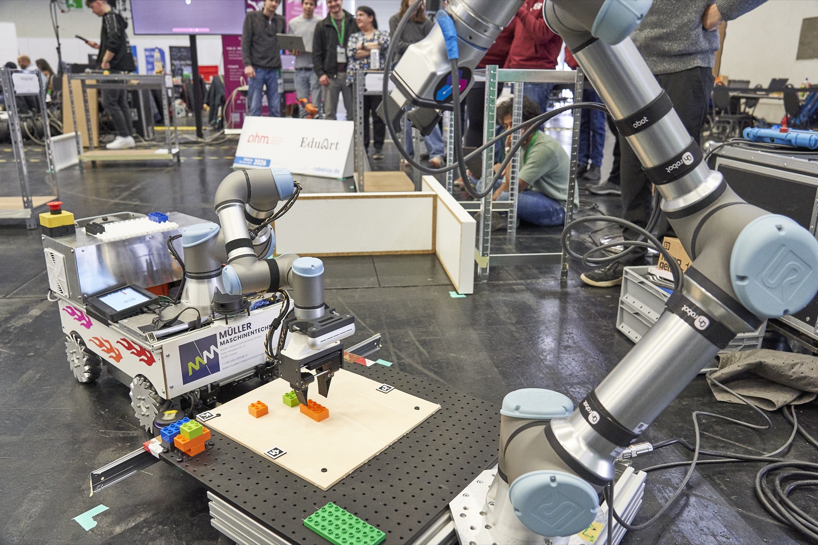 Der mobile Roboter montiert farbige Bauteile an einer Station, im Hintergrund Zuschauer und ein stationärer Roboterarm